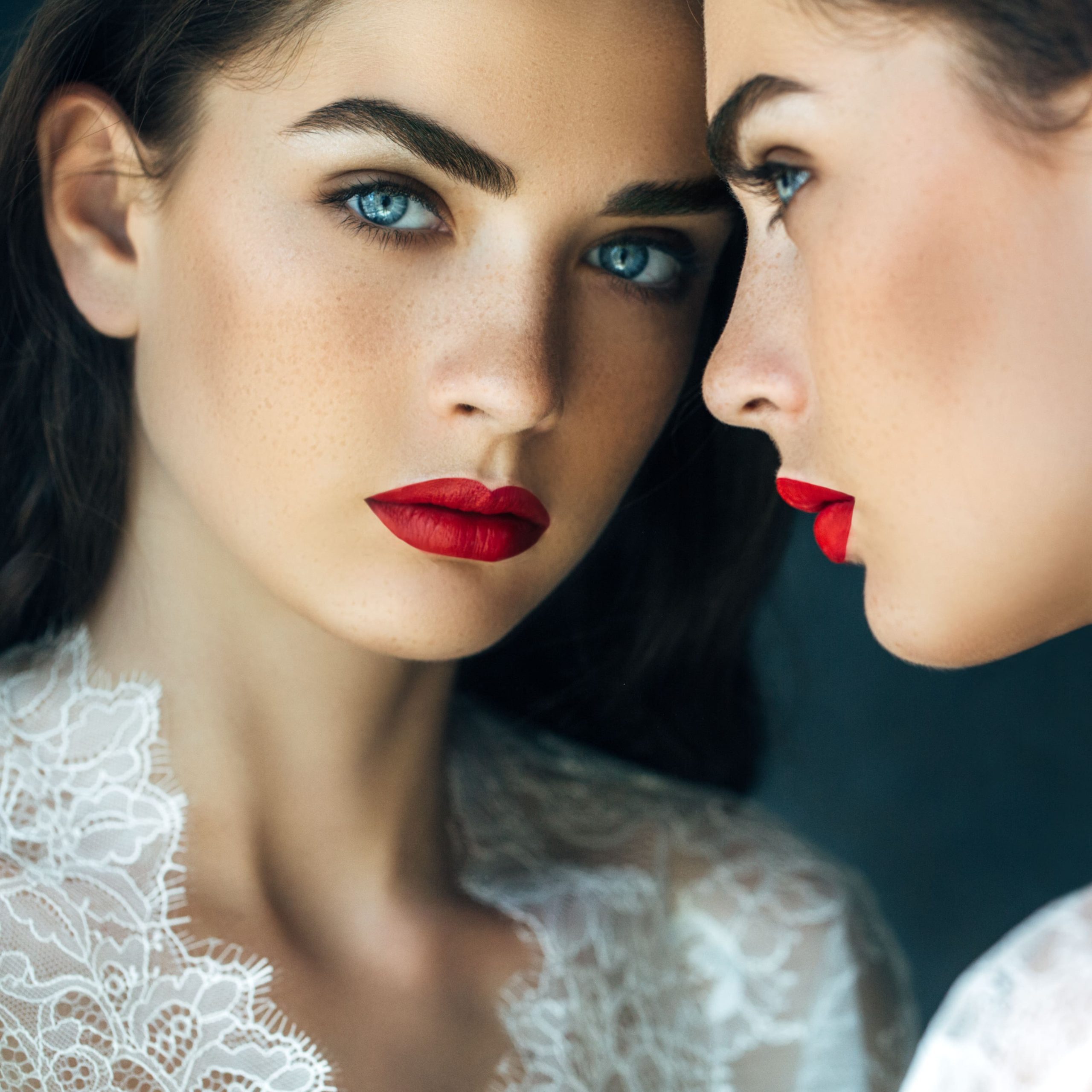 Two women with striking blue eyes and red lipstick facing each other, one with a lace top, in a close, reflective pose.